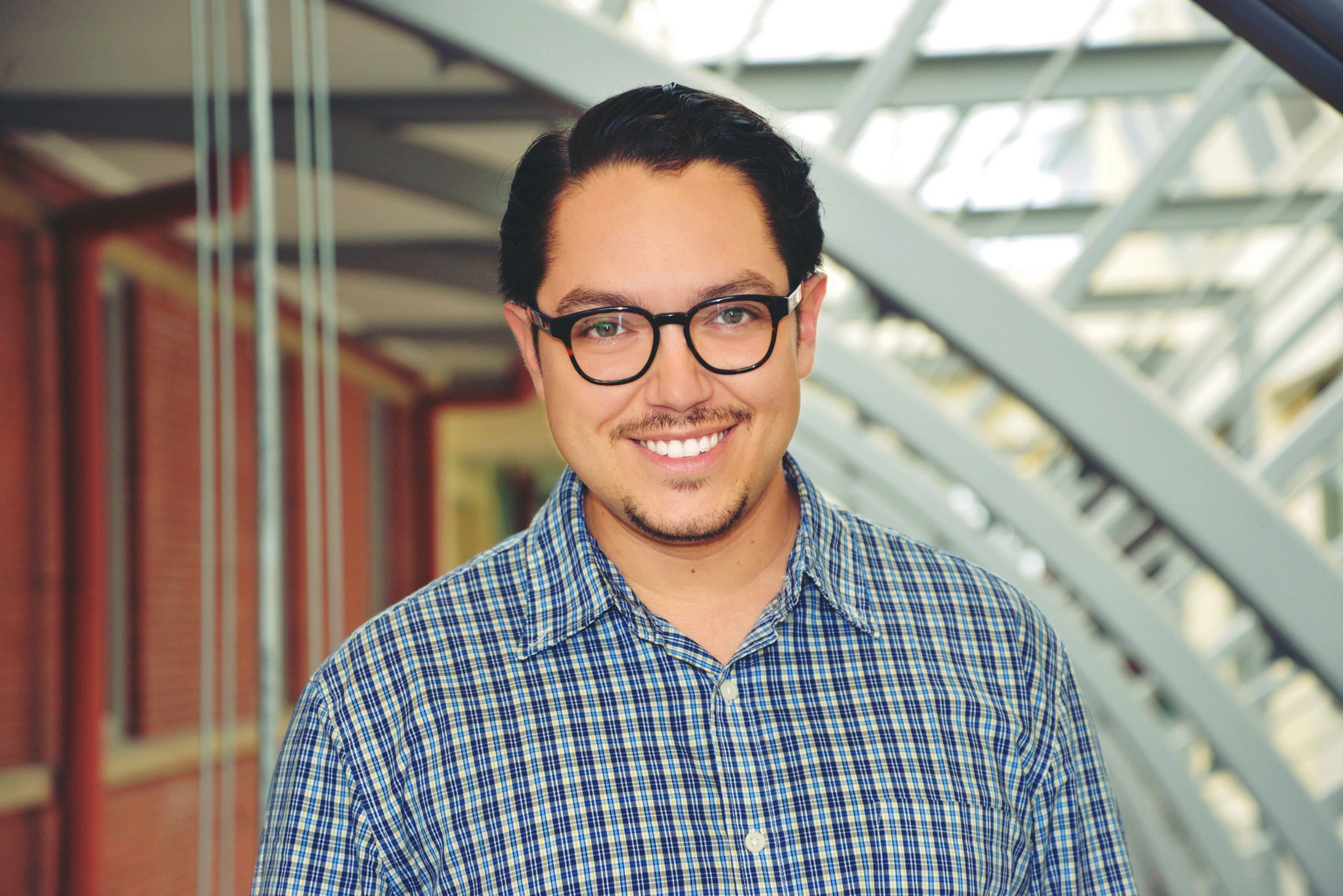 Oscar Ruiz (a Hispanic Latin-American man with short, dark brown hair and glasses, wearing a blue and white checked shirt), smiling into the camera.